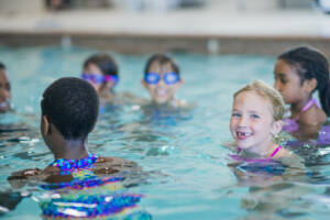 A small group of school-aged children are seen swimming in an indoor pool during a lesson. They are each wearing swimsuits and smiling as they tread water.