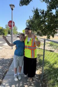 A crossing guard in bright yellow safety vest stands with a young student on a sidewalk near a stop sign, both waving at the camera.