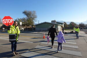 Crossing guard in yellow safety vest holds stop sign while people cross street at crosswalk on sunny day with mountains visible in background.